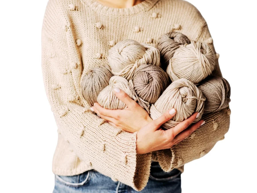 A person in a beige crochet sweater holds several skeins of neutral-colored yarn.