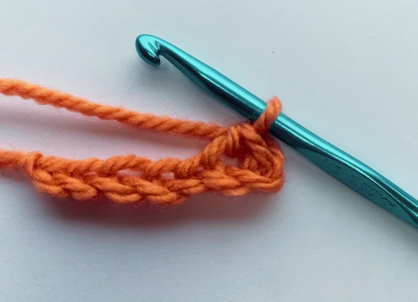 Blue crochet hook with an orange yarn chain stitch transitioning into a single crochet stitch on a white background.