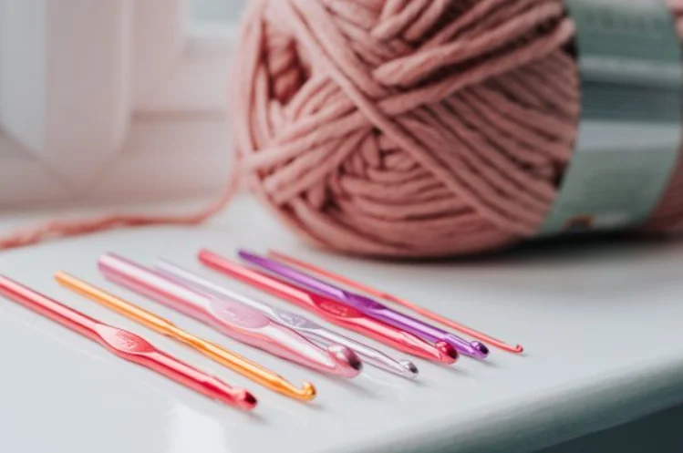 A set of colorful crochet hooks, ideal as crochet tools for beginners, is laid out on a white surface with a large ball of pink yarn in the background.