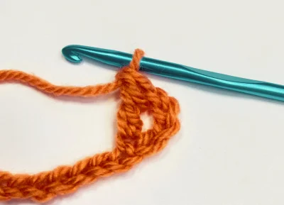 Close-up of a teal crochet hook with orange yarn forming the beginning chain stitch for a treble crochet stitch on a white background.