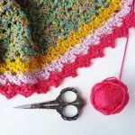 A colorful crocheted Spindle Shawl with pink, yellow, and multicolored yarn, a pink yarn ball, and ornate silver scissors arranged on a white background.