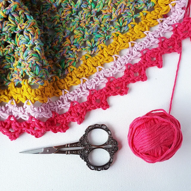 A colorful crocheted Spindle Shawl with pink, yellow, and multicolored yarn, a pink yarn ball, and ornate silver scissors arranged on a white background.