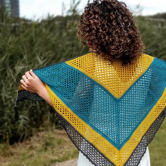 Person with curly hair displaying the Piper Shawl, a triangular crochet piece in blue, yellow, and black, outdoors with lush greenery in the background.