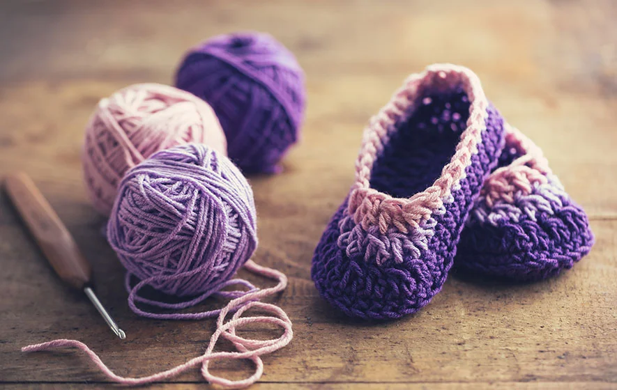 Crocheted purple and pink baby booties rest on a wooden surface, surrounded by three balls of yarn and a crochet hook. The detailed crochet stitches add an extra touch of charm to the cozy scene.