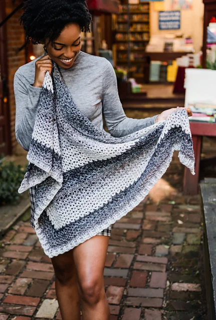 A woman wearing a gray long-sleeve shirt stands outside on a brick path, smiling and holding a large, striped knitted Omar Wrap shawl.