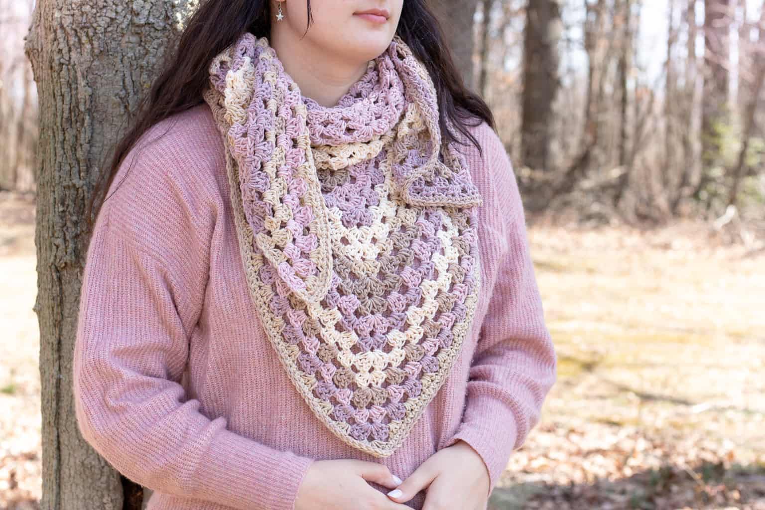 A woman wearing a light pink sweater and a crocheted triangle Spring Shawl in beige and lavender stands outdoors by a tree.