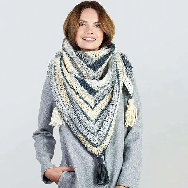 A woman wearing a grey coat and the Quick and Cozy Shawl—a large, multicolored, striped triangular wrap with tassels—stands against a plain light background.