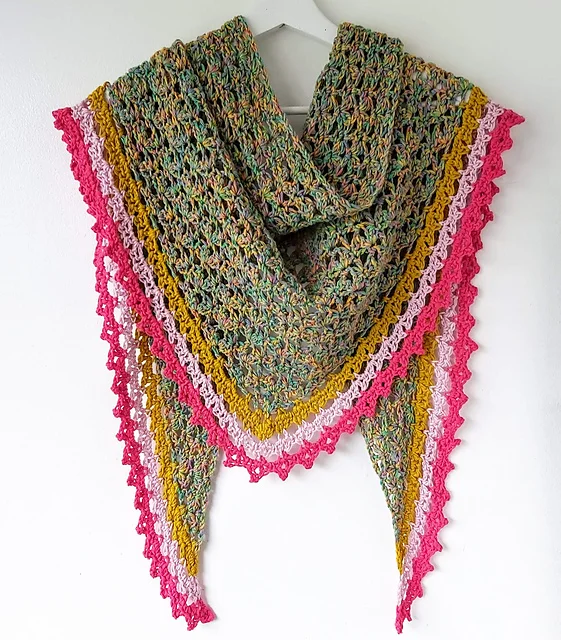 A multicolored Spindle Shawl with a lacy texture, featuring green and yellow tones and finished with pink and red scalloped edging, hangs on a white hanger.