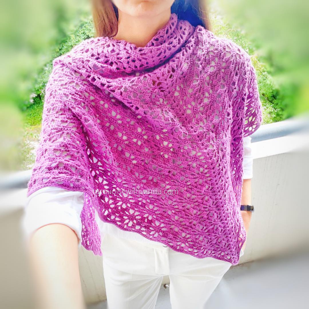 A person wearing a white outfit and a purple crocheted lace summer scarf stands on a balcony with greenery in the background.