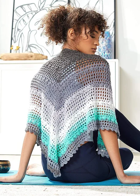 A woman sits on a mat wearing a Mesh and Blocks Shawl in shades of grey, white, turquoise, and teal. She is facing away, with art and decor visible in the background.