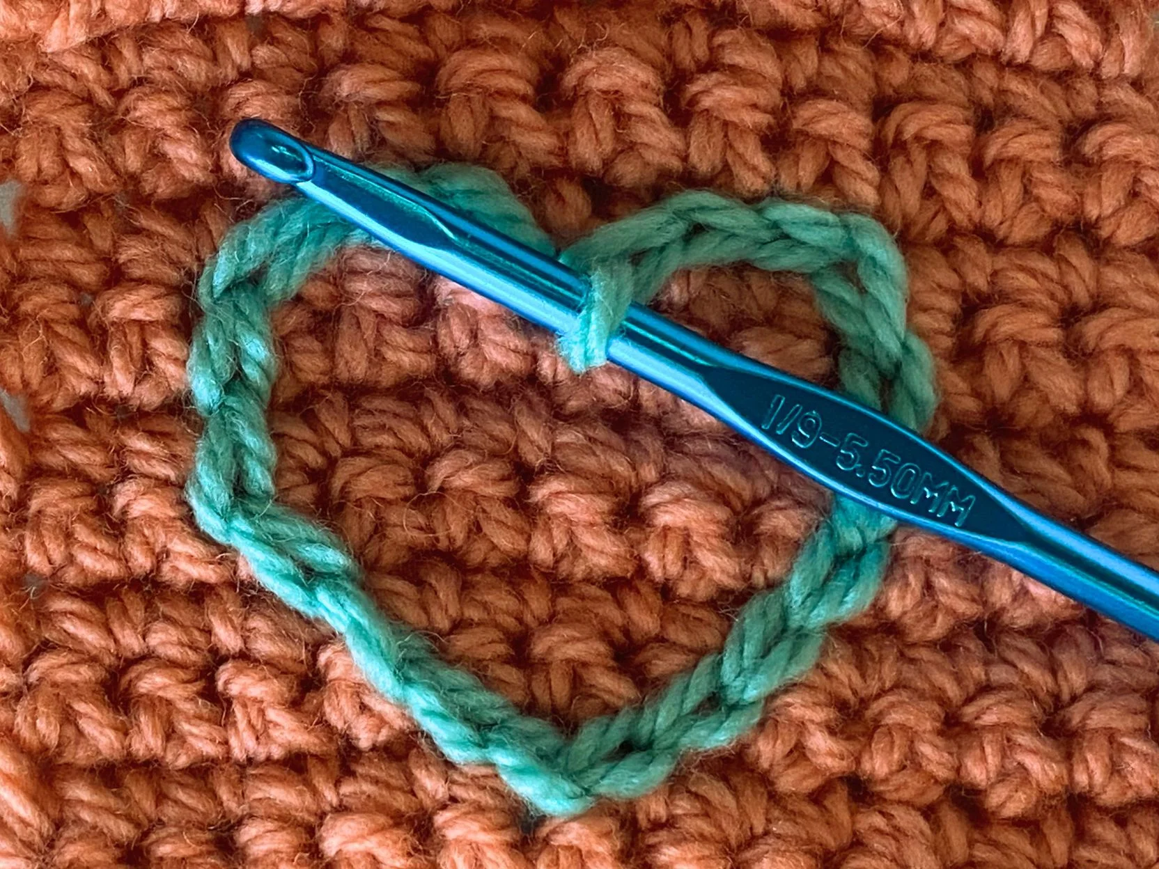 A blue crochet hook rests on orange crocheted fabric, while green yarn forms a heart, hinting at the delicate artistry of a slip stitch.