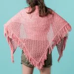 Person wearing a pink, fringed Summerday Shawl draped over their shoulders, standing against a blue background, facing away from the camera.