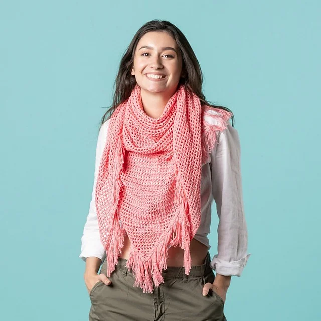 A woman stands against a light blue background, smiling and wearing a white shirt, green pants, and a large pink fringed Summerday Shawl.