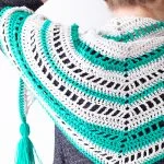 Person wearing a Stardew Triangle Scarf—a crocheted shawl with white, green, and black stripes and a green tassel—photographed from the back and side.