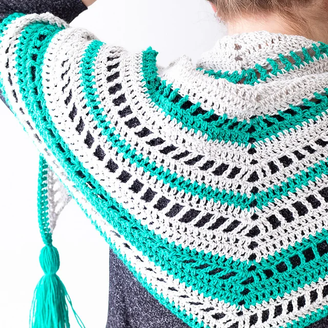 Person wearing a Stardew Triangle Scarf—a crocheted shawl with white, green, and black stripes and a green tassel—photographed from the back and side.