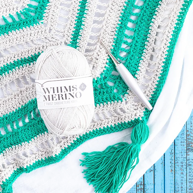 A ball of Whims Merino yarn, a crochet hook, and a green and white Stardew Triangle Scarf with a tassel are displayed on a blue wooden surface.