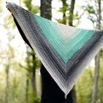 A person holds up a large, triangular crochet Beginner One Skein Shawl with gradient colors from grey to teal, standing outdoors in a wooded area.