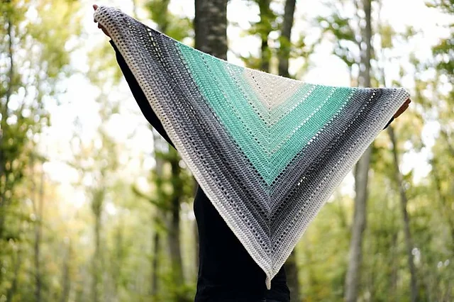 A person holds up a large, triangular crochet Beginner One Skein Shawl with gradient colors from grey to teal, standing outdoors in a wooded area.