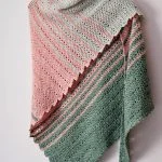 A crocheted Willow Shawl in pastel green, pink, and gray shades hangs on a wooden hanger against a white wall.
