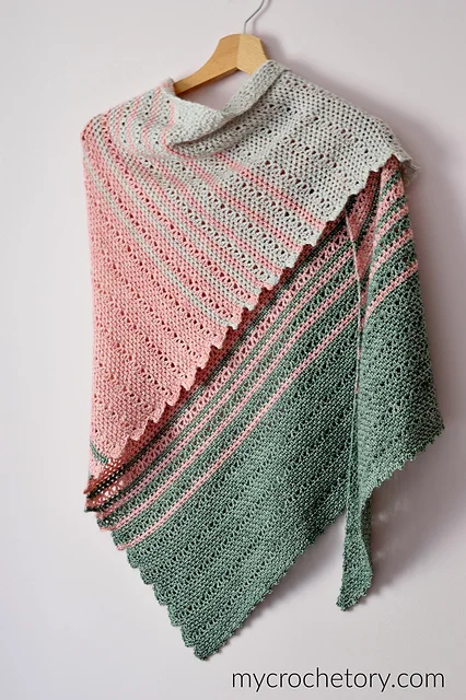 A crocheted Willow Shawl in pastel green, pink, and gray shades hangs on a wooden hanger against a white wall.