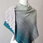 The Willow Shawl is a knitted shawl with a gradient color transition from light gray to teal, beautifully displayed on a white mannequin torso.