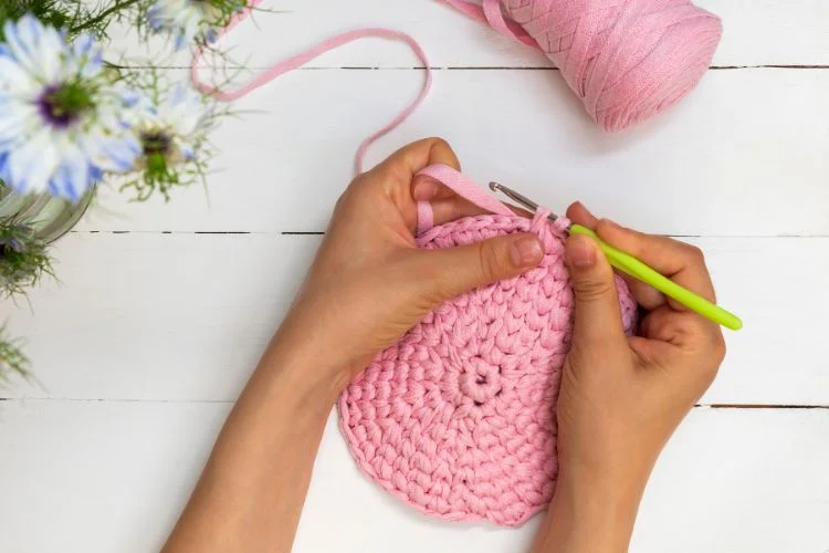 A woman is crocheting a pink crocheted bag on a wooden table, demonstrating how to crochet a circle.