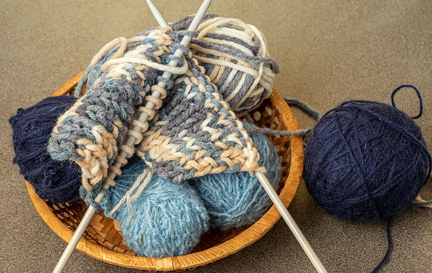 A knitting project featuring blue and beige yarn rests in a basket, with needles poised for more rows of stockinette stitch. Two balls of yarn lie beside it on a textured surface, ready to weave their colors into the growing creation.