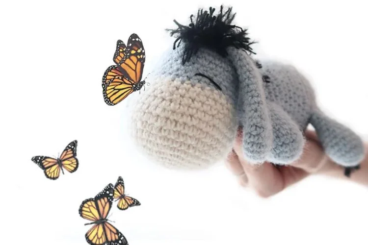 A person is holding a crocheted donkey with butterflies flying around it.