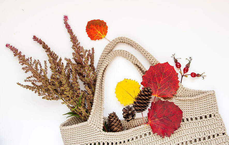 A crochet tote bag holds dried branches, pinecones, and colorful autumn leaves against a white background.