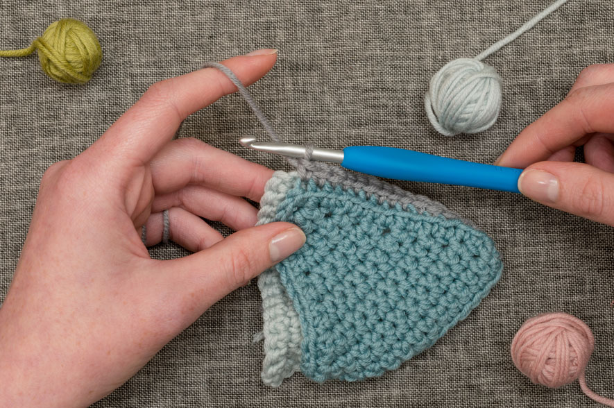 Hands skillfully engaged in corner to corner crochet, crafting a blue and gray piece with a blue-handled crochet hook against a gray fabric background, surrounded by small balls of yarn.