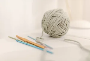 A ball of light gray cotton yarn for crochet sits beside three crochet hooks on a white surface.