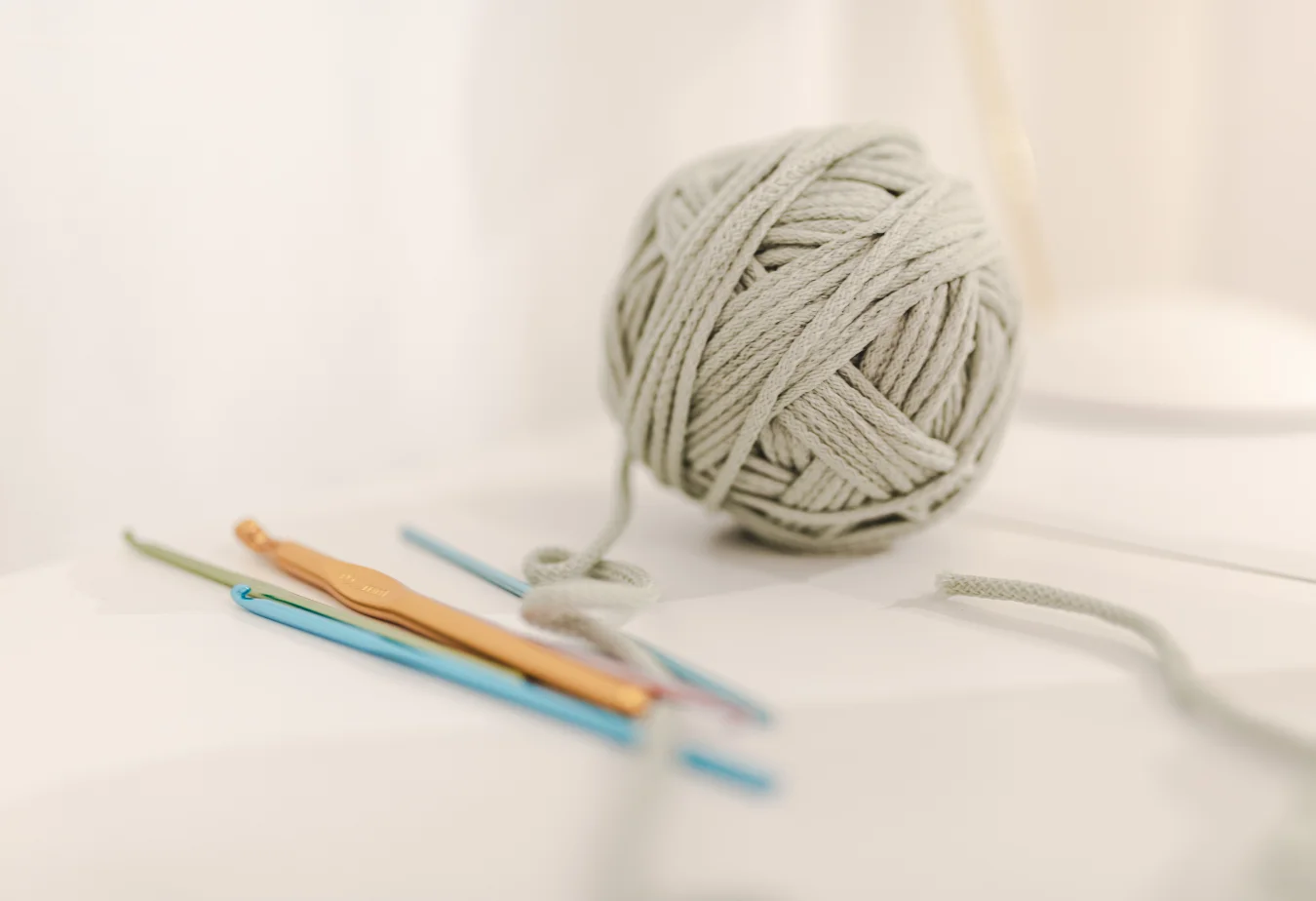 A ball of light gray cotton yarn for crochet sits beside three crochet hooks on a white surface.