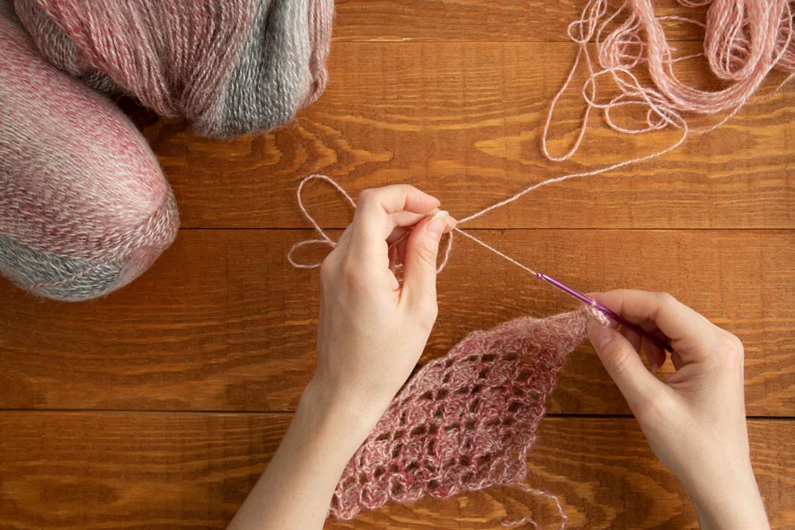 Hands carefully crocheting with pink yarn on a wooden table, a reminder that even crochet mistakes can lead to unexpected beauty.