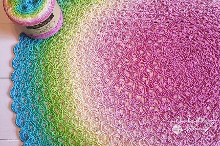 A rainbow colored crocheted shawl with a ball of yarn next to it.