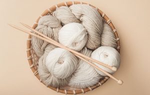 A basket filled with beige and cream-colored worsted weight yarn balls, accompanied by two wooden knitting needles.