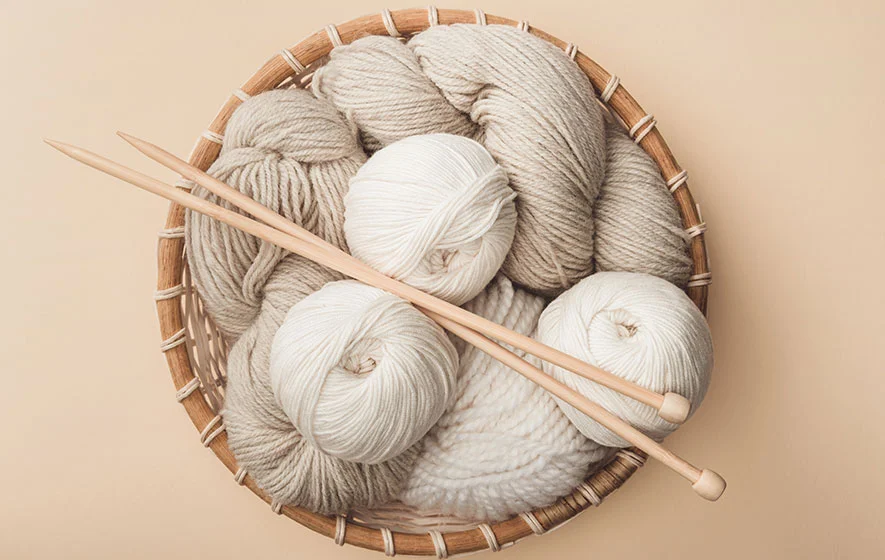 A basket filled with beige and cream-colored worsted weight yarn balls, accompanied by two wooden knitting needles.