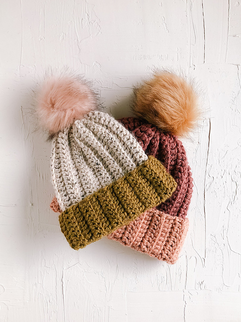 Two cozy 1.5 Hour Beanies with large pom-poms—one in cream and olive green, the other in rust and pink—are placed side by side on a white textured surface.