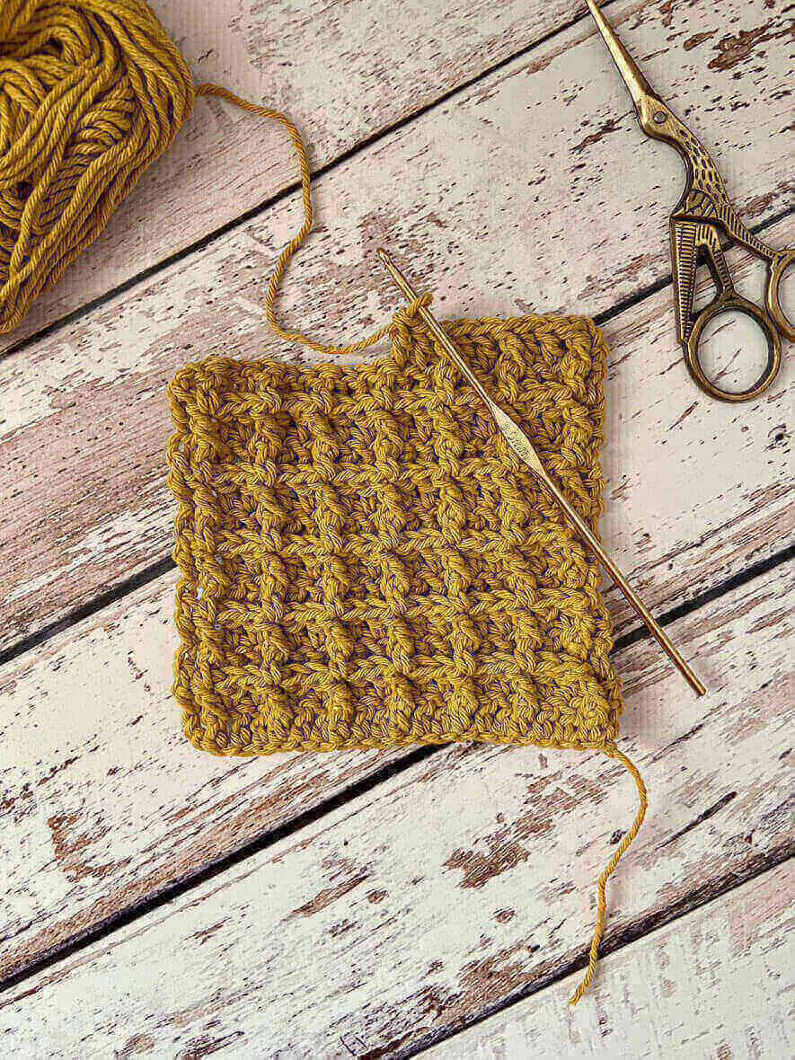 A square piece of yellow crochet waffle stitch rests on a wooden surface, accompanied by a crochet hook. Nearby, scissors and a ball of yarn complete the cozy scene.