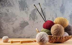 A basket brimming with various types of yarn, showcasing an array of colors, alongside knitting needles on a wooden surface, set against a textured gray background.