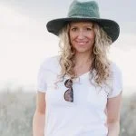 A woman with long curly hair wearing a green Cleo Wide Brim Fedora, white t-shirt, and jeans stands outdoors with sunglasses hanging from her shirt pocket.