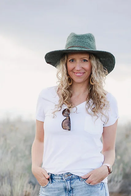 A woman with long curly hair wearing a green Cleo Wide Brim Fedora, white t-shirt, and jeans stands outdoors with sunglasses hanging from her shirt pocket.