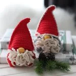 Two crocheted Christmas Gnome figures with red hats and white beards sit on wrapped gifts, nestled among pinecones and evergreen branches.