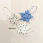 Three crocheted Snowflake Ornaments, two white and one blue, with looped strings, are displayed on a light wooden surface.
