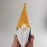 A hand holds a small crocheted Mini Norwegian Nisse Christmas Decoration, featuring a tall yellow hat, white beard, and round nose against a plain light background.