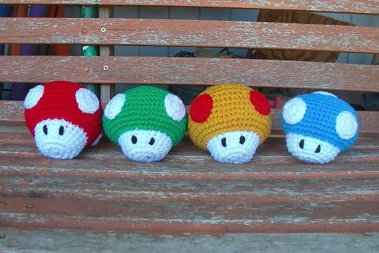 Four crocheted nintendo mario amigurumi dolls sitting on a bench.