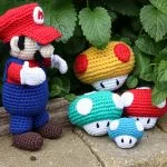A crocheted Mario figure stands on bricks next to colorful Mario Mushrooms in red, blue, and yellow, with green plants in the background.