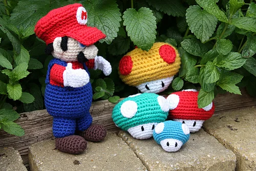 A crocheted Mario figure stands on bricks next to colorful Mario Mushrooms in red, blue, and yellow, with green plants in the background.