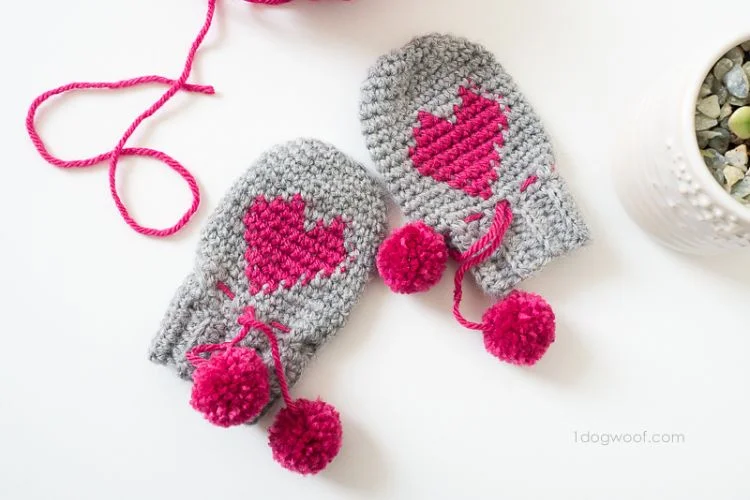 Two crocheted mittens with pink pom poms.