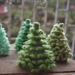 Three small, green, crocheted Christmas trees are displayed on a wooden surface, their charming Christmas tree shapes standing out against a blurred outdoor background.