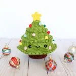 A festive Christmas Tree Ami, crocheted with colorful pom-poms as ornaments and a yellow star on top, is surrounded by four decorative baubles on a light wooden surface.